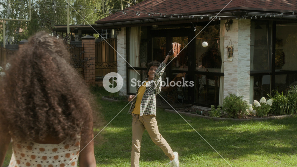 Medium full slowmo shot of cheerful teenage African American girl tossing ball up in air, then throwing to brother with baseball glove, boy missing and both laughing, while having fun outdoors on lawn