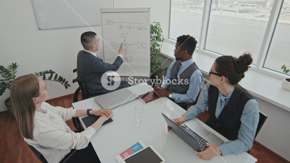 Handheld high angle shot of disabled manager in wheelchair pointing at whiteboard and delivering business strategy presentation to team of colleagues in meeting room