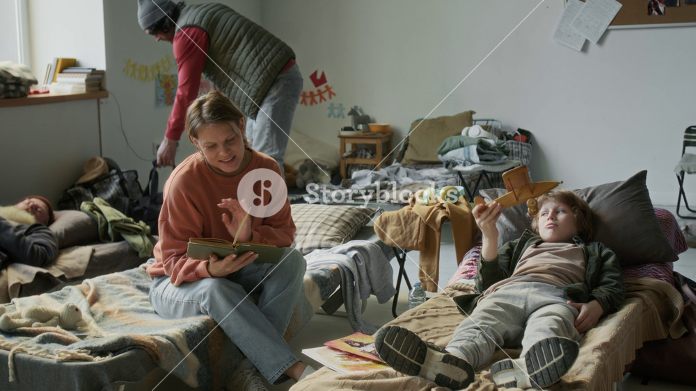 Full shot of young family staying in refugee camp during natural disaster, mother reading aloud book while son is playing with toy airplane, senior man sleeping, guy carrying bag walking in