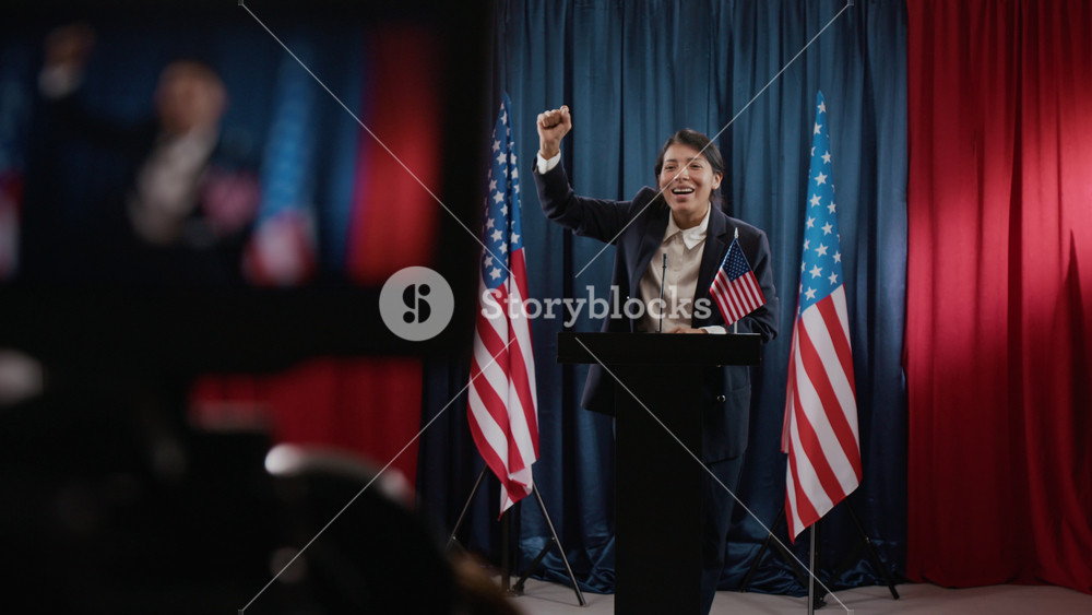 Camera filming young Hispanic woman politician standing at podium raising fist in victory during speech with USA flags in background. Clip is ideal for political campaigns and election news