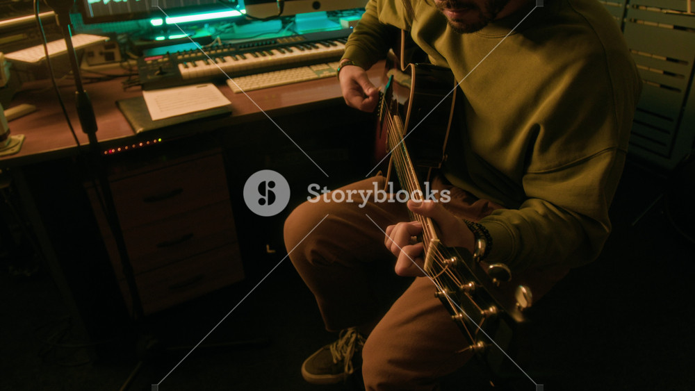 Side tilt shot of Caucasian male artist playing guitar in modern recording studio, recording sound of instrument for new song or podcast, audio sequences on computer screens in background