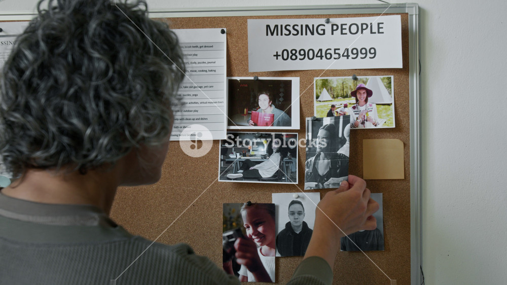 Rear medium closeup of unrecognizable mature woman with curly graying hair standing in front of information board on wall in refugee facility, looking at photos of missing victims of natural disaster