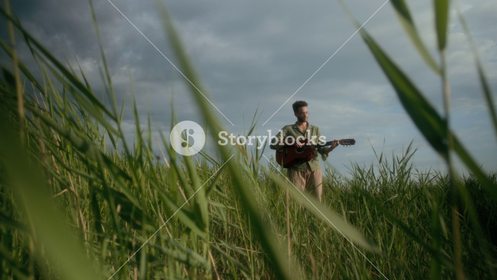 Long shot of lonely young male musician standing in field in nature, against cloudy sky, playing sad song on guitar, filmed from beneath green grass