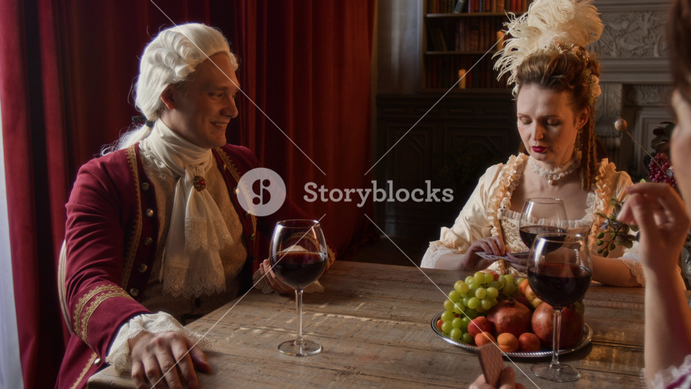 Medium shot of noble Victorian period gentleman in red surcoat and wig and two middle-aged lady friends sitting around table in palace at daytime, enjoying red wine and playing card game