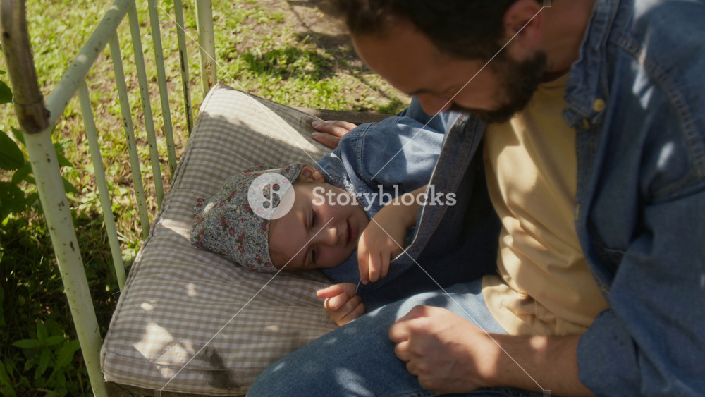 Medium high-angle shot of rural Caucasian man putting little daughter to sleep for midday nap outdoors in fresh air in garden on summer day, dreamy girl talking to father, family bond and loving care
