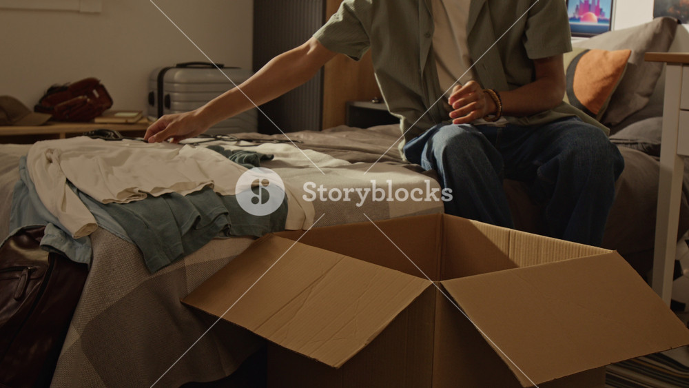 Handheld shot of Black male teenager folding clothes and packing cardboard box in bedroom, preparing for relocation. Footage is ideal for moving services, student housing, or decluttering concepts