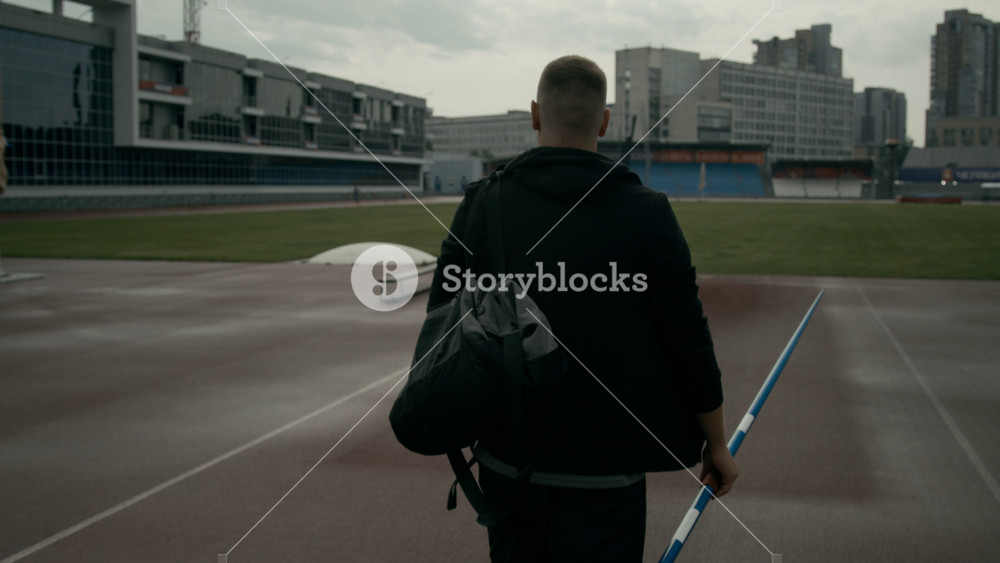 Medium rear follow shot of unrecognizable male athlete in black shorts and sweatshirt walking across stadium field, carrying javelin and bag, on way to training session