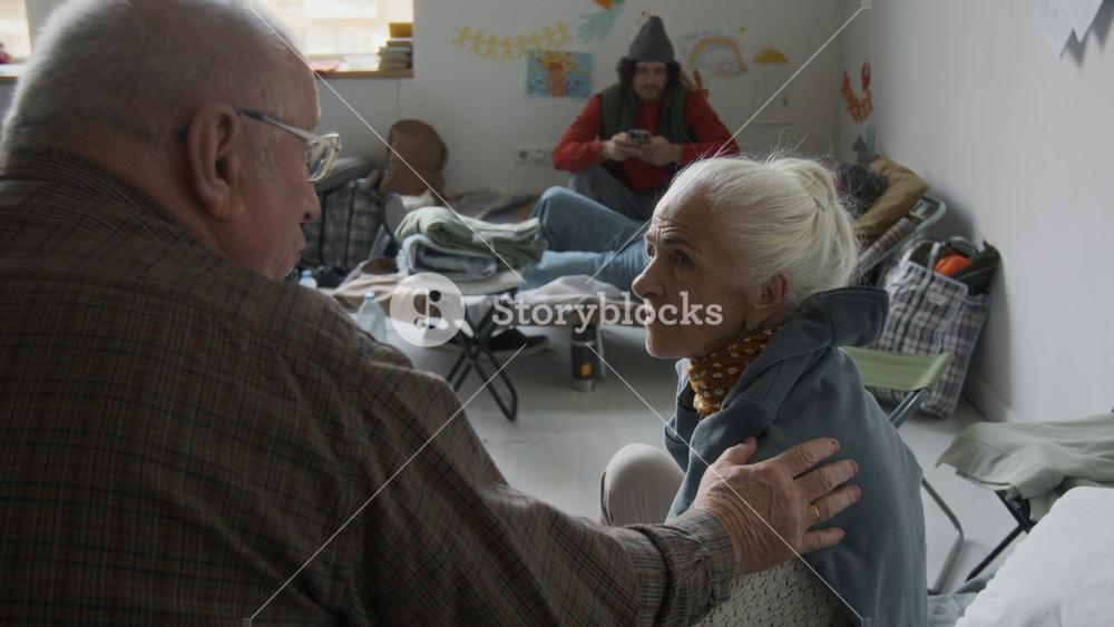 Medium rear shot of elderly Caucasian husband and wife sitting on makeshift bed in refugee shelter, surviving flood or fire, discussing their trauma, talking and comforting each other