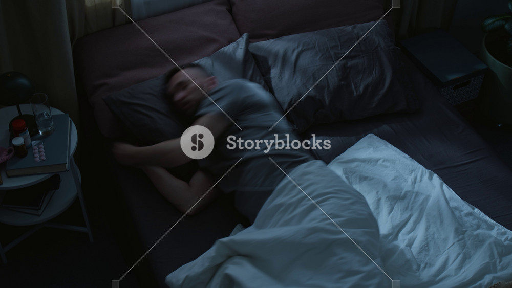 Top view time lapse of young Caucasian man in pajama pants and t-shirt sleeping in bed at night in dark bedroom, tossing and turning, hugging pillow, covering himself with blanket, talking in dream