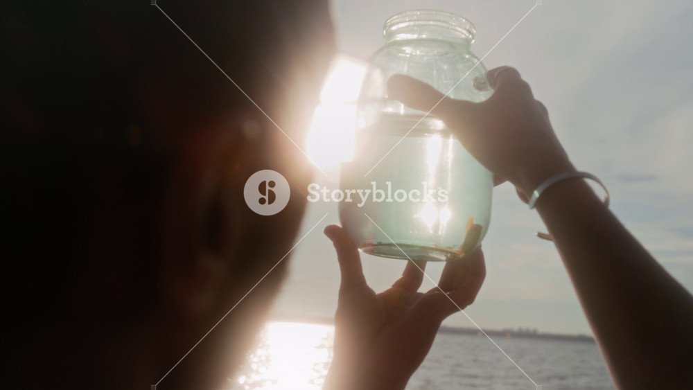 Rear over-shoulder closeup of hands of black man in glasses holding up jar with captive or pet goldfish against bright sun while relaxing on lake beach in afternoon