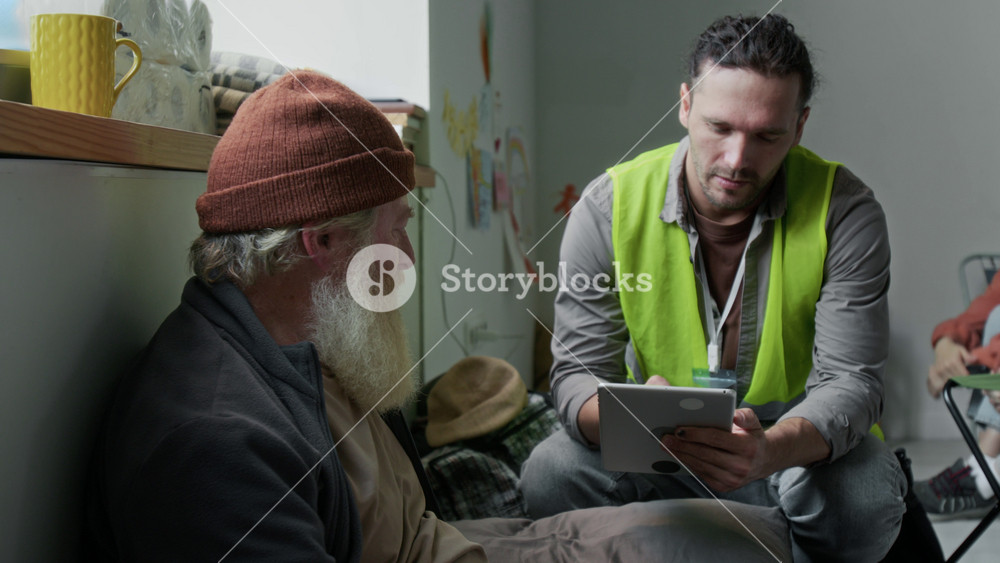 Medium full shot of cheerful Caucasian male social worker in yellow vest sitting on chair next to elderly displaced man with beard, asking questions, filling form, while responding to natural disaster