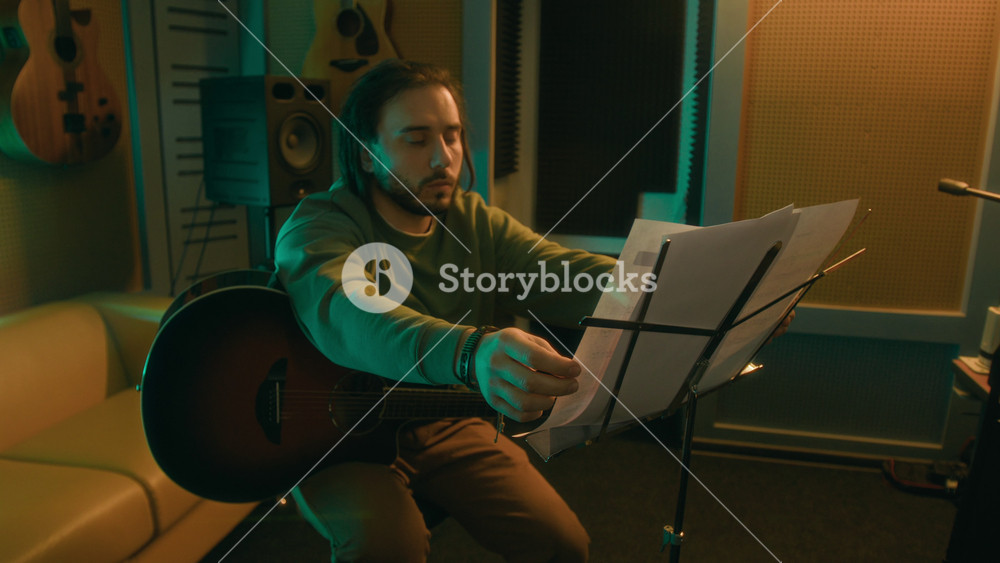 Medium long shot of young adult Caucasian man with guitar sitting on chair and placing sheet music on music stand in neon lit studio, studying notes