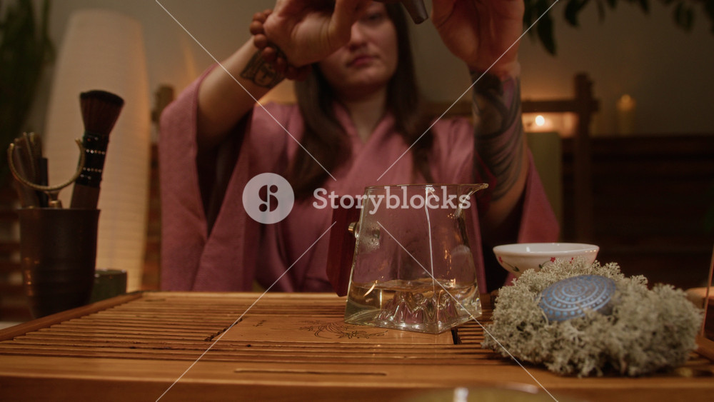 Medium shot of calm, peaceful young female gongfu master in pink silk kimono, with tattoo on arm pouring hot water in glass jug after warming up clay pot with graceful, practiced movements
