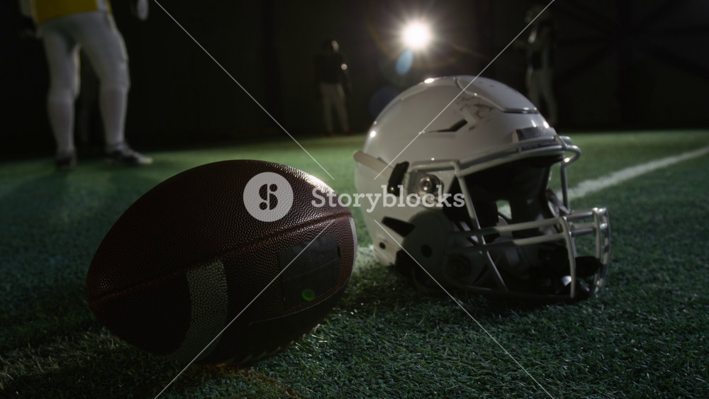 Close-up of American football protective helmet with face mask and leather ball lying on green turf of dark gridiron arena during team practice session, group of anonymous male players training