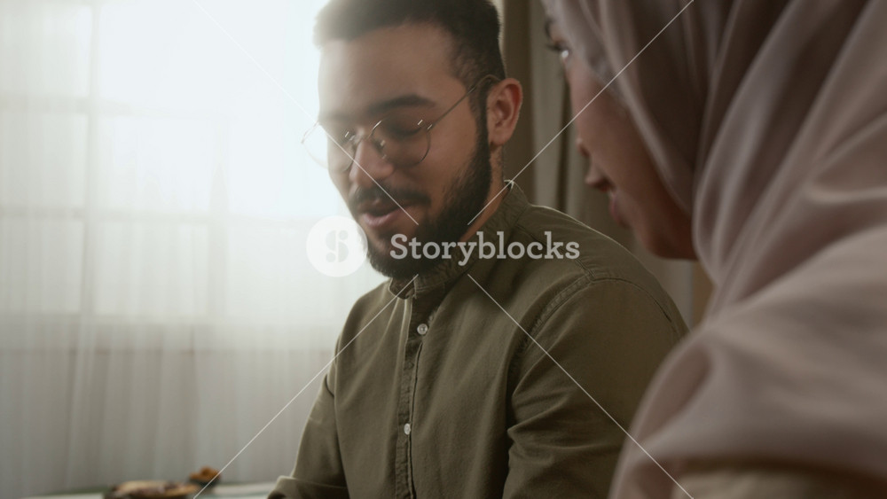 Cinematic tilt up shot of young Muslim couple reading Quran together at home with focus on smiling young man discussing verses with wife, copy space