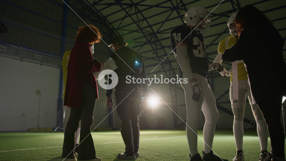 Full low angle backlit shot of TV cameraman talking to gridiron football team captain about shoot plan, female producer running up with storyboard while stylist checking players uniforms and gear