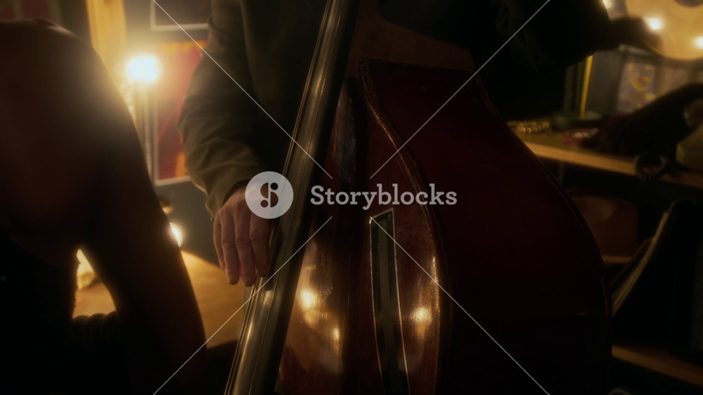 Pan down shot of adult Caucasian man wearing suit and hat playing upright bass backstage in jazz club, hands strumming strings