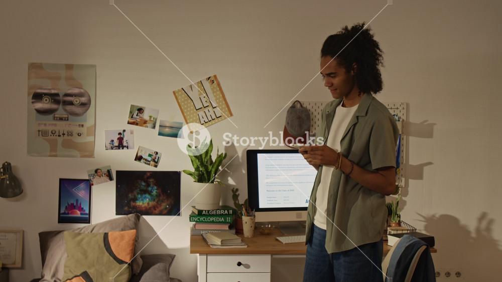 Handheld shot of African American teenager celebrating college acceptance on computer screen, dancing and talking on smartphone. For higher education marketing and student success stories