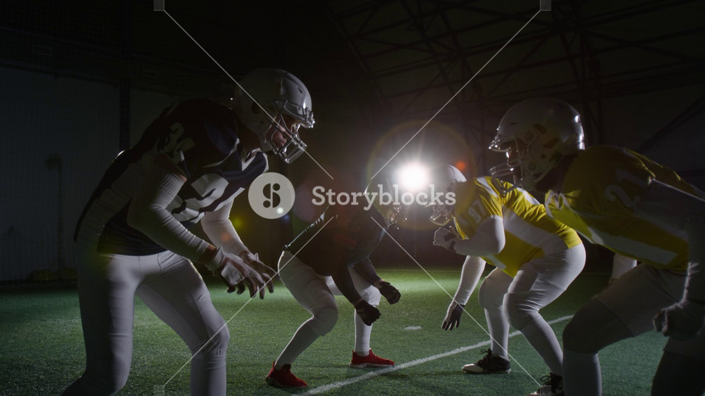 Full side shot of rival American football athletes from opposing teams, wearing yellow and blue jerseys and protective gear, crouching against each other on scrimmage line at sports arena