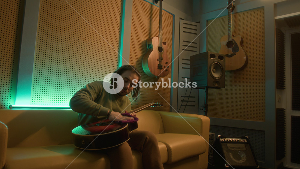 Medium long shot of male young adult guitarist sitting on couch and wiping finger board and body of acoustic guitar in neon lit recording studio, taking care of string instrument