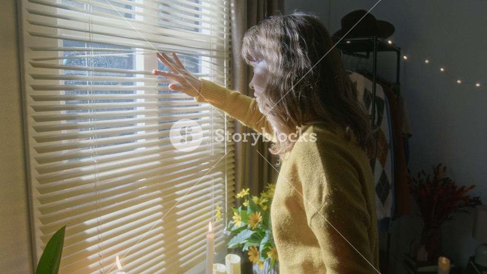Caucasian young woman sitting on bed, closing book, walking to window and peeking through blinds. Video suitable for campaigns about home security, loneliness, or neighborhood watch