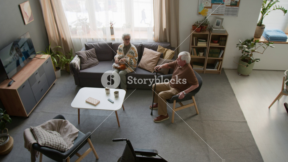 Wide angle top view shot of seniors in retirement home common room, playing chess and knitting, senior man walking with cane to sit in armchair. Ideal for senior care and assisted living
