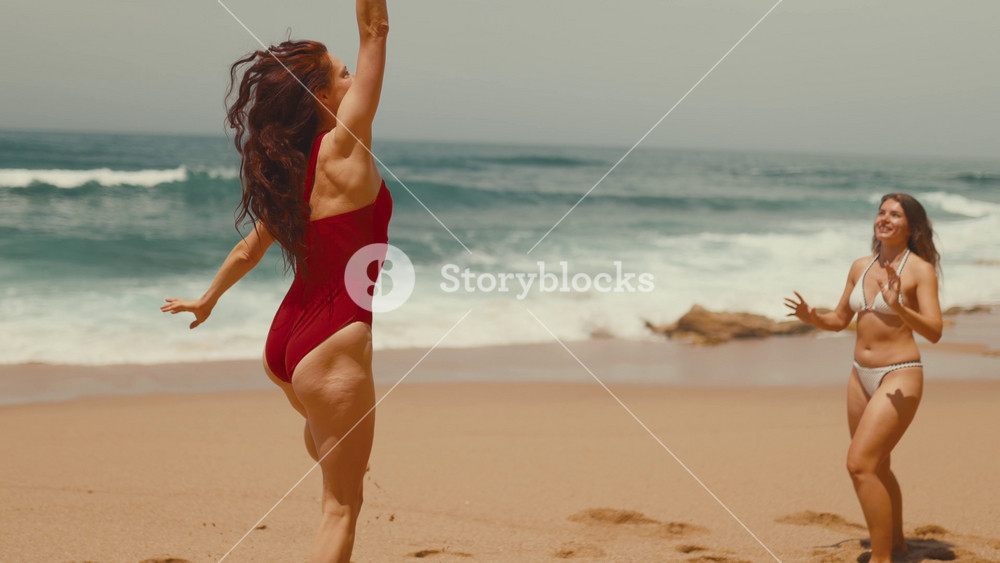 Caucasian female beachgoer in red swimsuit plays with colorful beach ball, running along sandy shore with ocean waves and another woman in