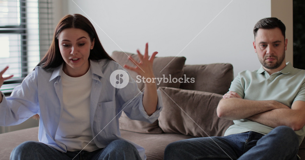 Couple sitting on sofa in living room arguing over disagreement, woman yelling at man, man experiencing psychological abuse from his wife