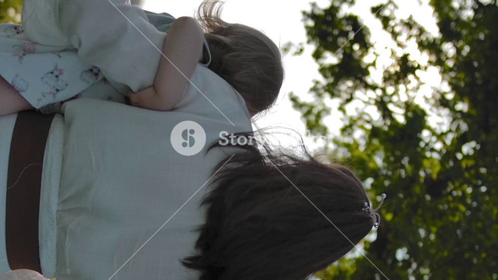 Mom in a white dress with daughter in her arms goes to an old oak tree with a swing in the middle of a green field. Slow motion. Sun glint makes flare of the sun through the foliage. Back view. Vertical shot.