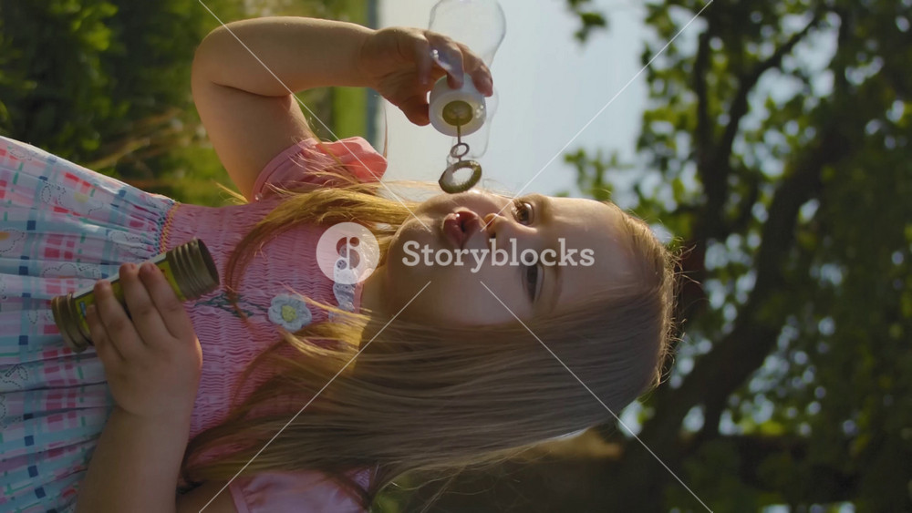 Front view of little girl in blows soap bubbles. Close-up of a happy child. Healthy lifestyle for children in the fresh air outdoor. Far from the city. Slow motion. Sunset warm lighting. Vertical shot.