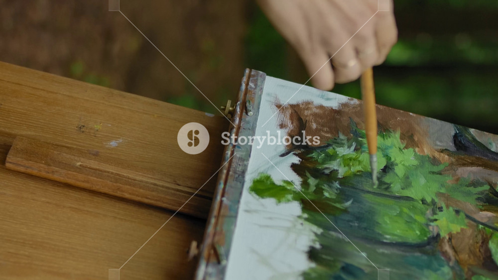 The girl's hand makes a few strokes on the landscape drawings in the forest. Favorite hobby in the fresh air. Frames wooden easel brush hand holding earth and trees. Middle shot. Vertical shot.