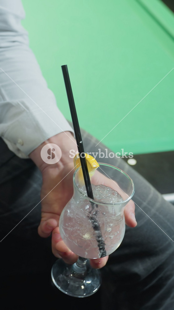 Close-up of young man wearing white shirt and grey trousers, sitting on green pool table holding cue stick and glass of lemon drink with