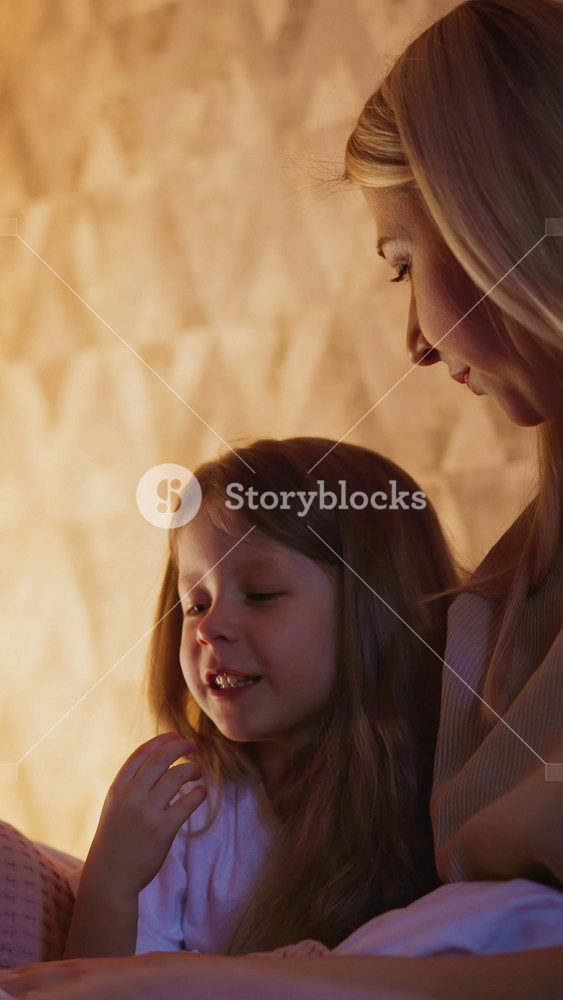 Happy little long haired girl reads diligently book sitting with careful mum on bed near play wigwam in partially illuminated bedroom close