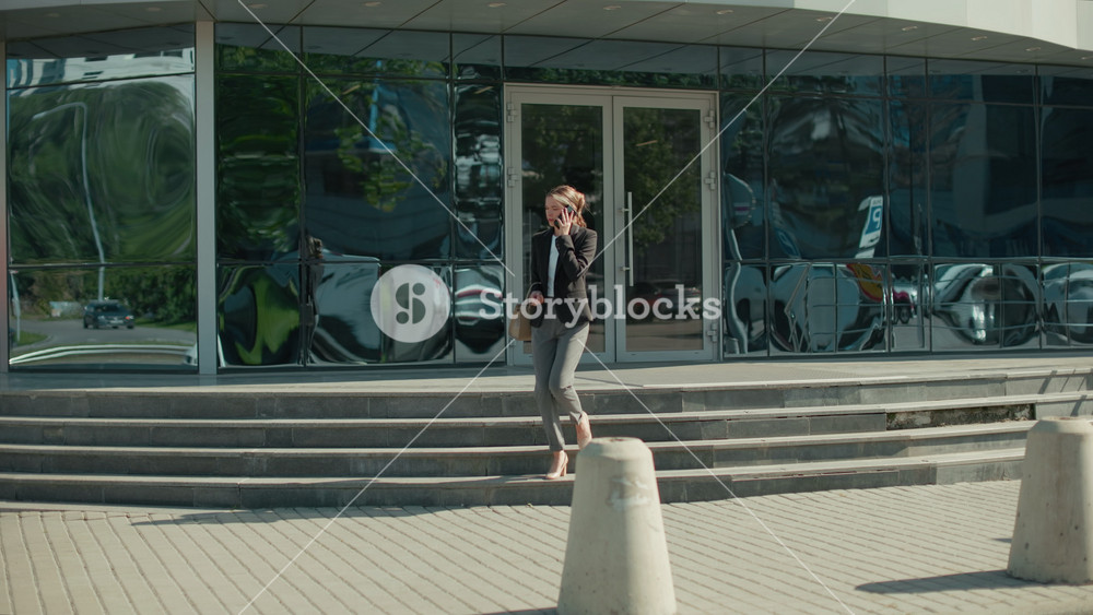 Banker on phone call stepping down staircase outside modern office building, holding handbag with sleek reflections of cars and surroundings