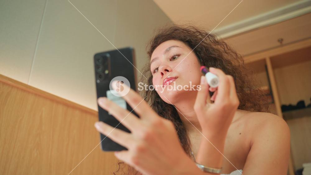 Hotel vanity evening woman applies cream in mirror with phone, soft warm lighting, closeup of delicate eye area, bracelet on wrist