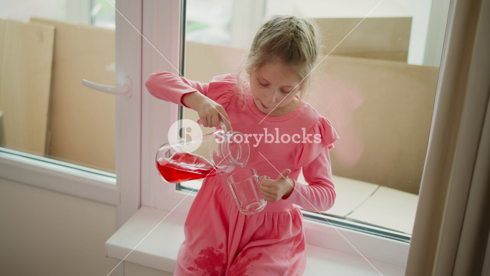 aerial view of kid wearing pink dress seated by window pouring red juice from jar into clear mug spilling drink on cloth indoors playful