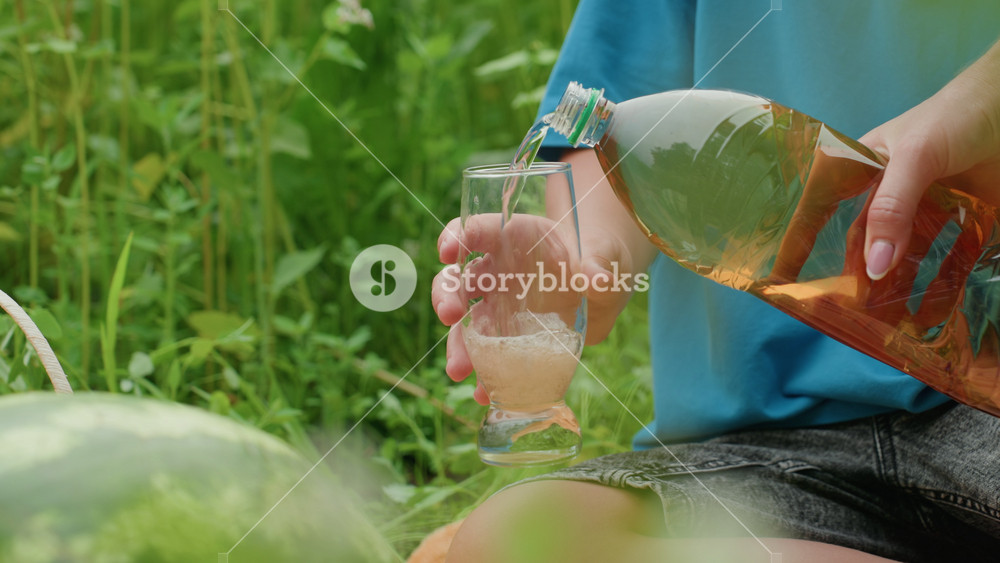 white person pouring bottle into glass on picnic blanket by tall grass with watermelon in foreground, closeup of hand and bracelet as golden