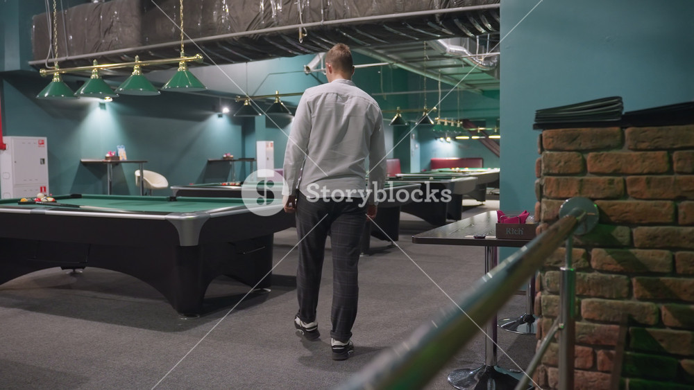 ack shot of player in white shirt and grey trousers walking into billiard room, standing in front of green pool table, pick up white ball