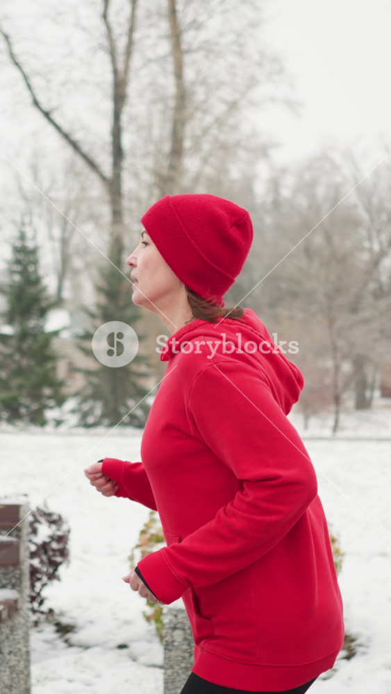 side view of tired lady jogging along snowy park pathway in red hoodie and black leggings surrounded by benches colorful bushes frosty