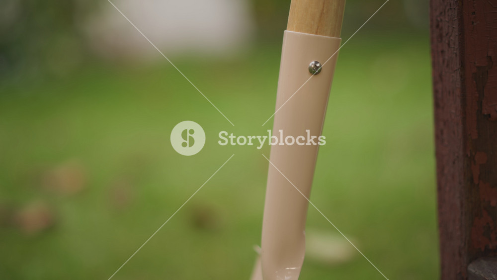 Closeup shovel shaft leaning fence post soft green lawn bokeh, metal ferrule, wooden texture with subtle raindrops, autumn leaves out of