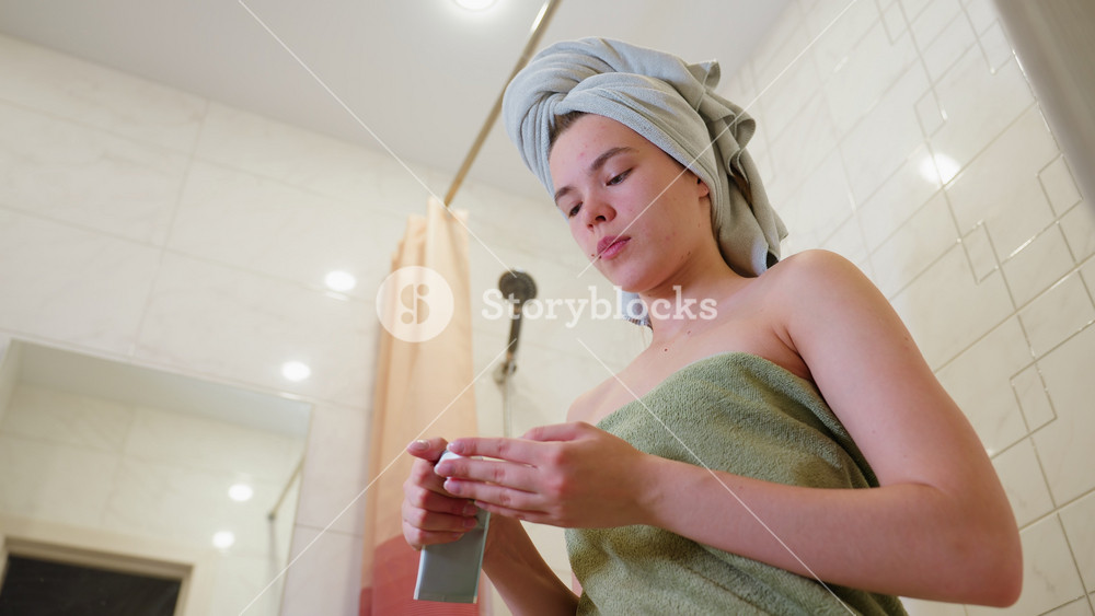 Close-up view of woman wrapped in towel gently applying cream on neck in bathroom after shower, highlighting soothing skincare moment with