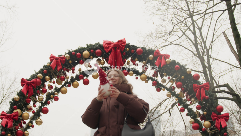 White woman under decorated arch, holding red gnome and looking up at garland and ribbons, bundled in warm coat, tourist vibe on winter