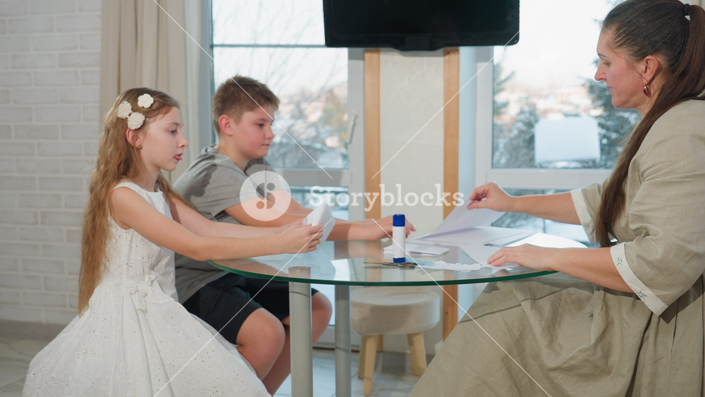 Side view of boy in gray top, black shorts, girl in white gown, and home teacher in gray gown sitting together creating craft with white