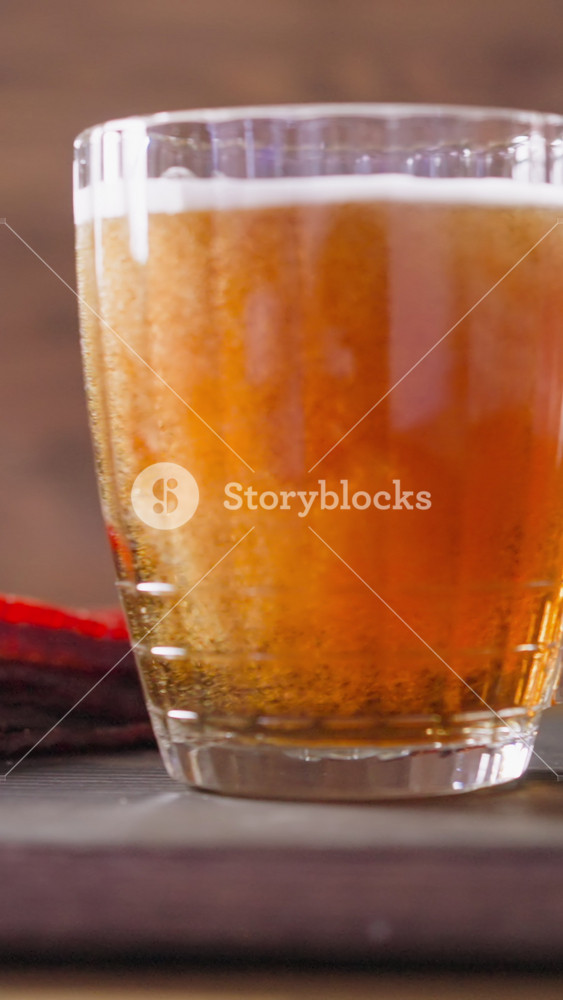 Heap of sun-dried salmon sticks and glass mug of fresh beer with rising up bubbles on rustic wooden cutting board in pub closeup slow motion