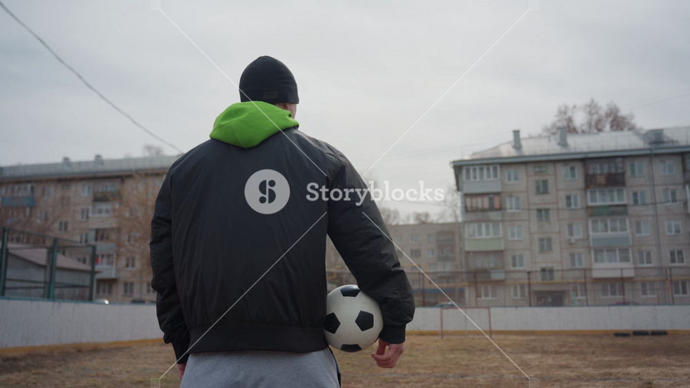 male strolls confidently, man with football outdoors, young man carrying soccer ball in muted weather, person dressed in hoodie