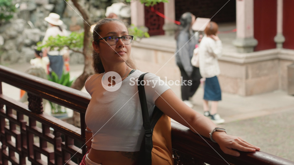 Young woman leaning on railing temple courtyard with carved wooden balustrade and rock garden casual traveler wearing glasses and backpack