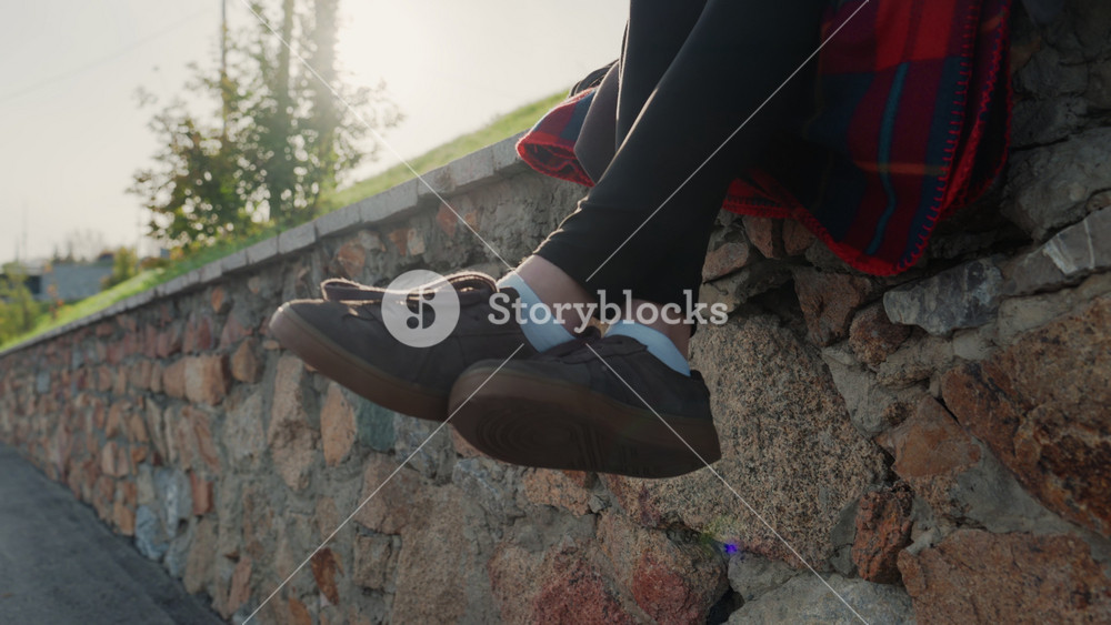 Golden light shoes on stone wall, late afternoon sun rimlighting legs, red plaid coat edge, brown sneakers, white socks, calm suburban lane