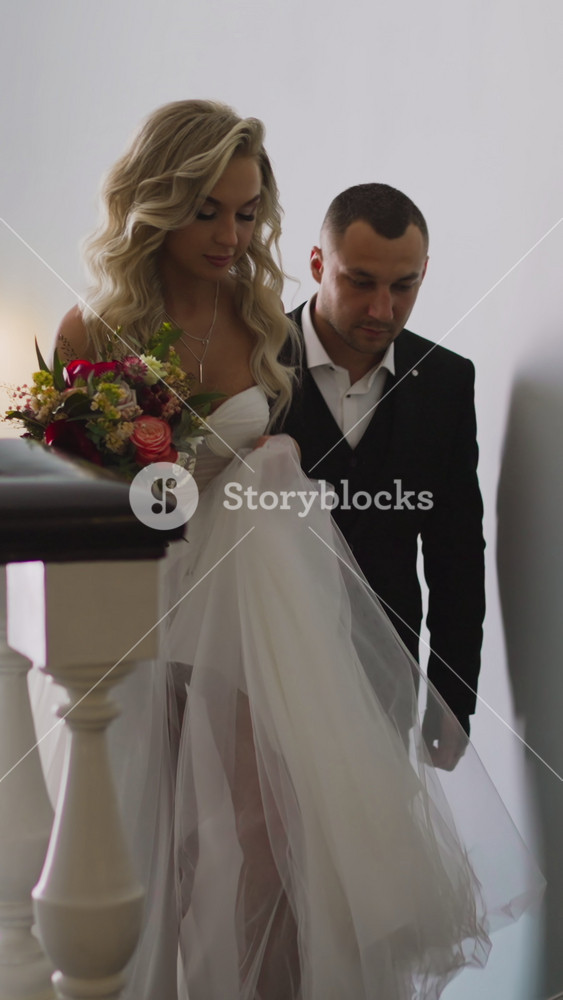 Pretty woman in wedding dress with hairstyle holding festive bouquet and groom in classic suit go up stairs in light hotel building hallway