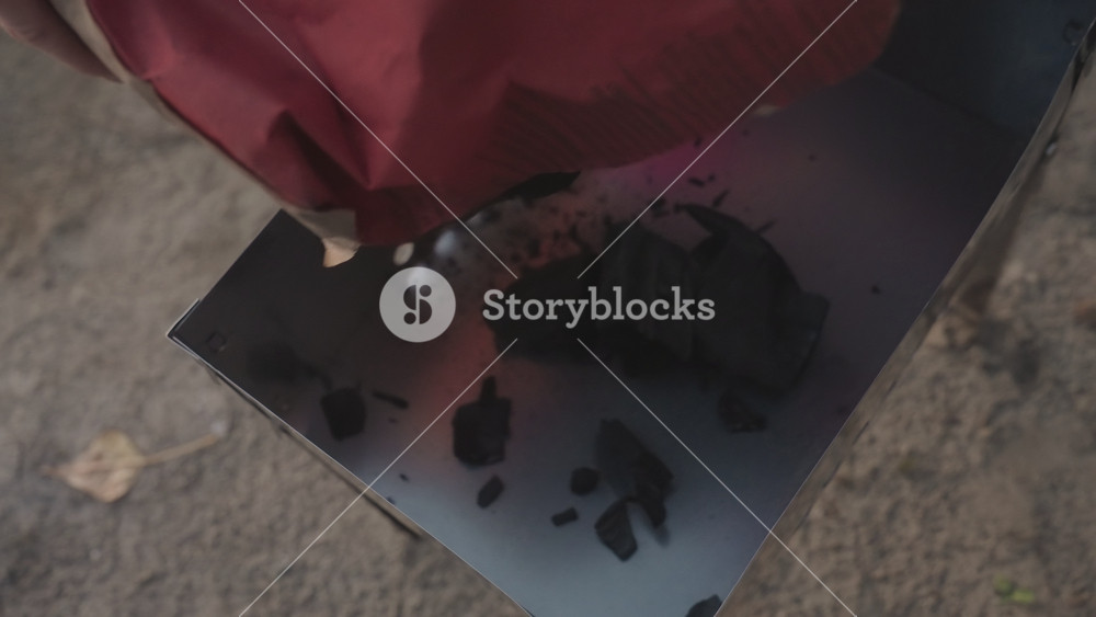 Camping charcoal poured into portable grill, red bag tipping briquettes onto metal tray on dirt, closeup of dark coal with smoke and ash