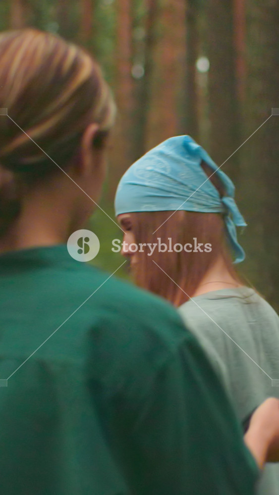 Back view of young woman in green shirt helping friend with blue bandana to put on backpack in peaceful forest, sunlight filters warmly
